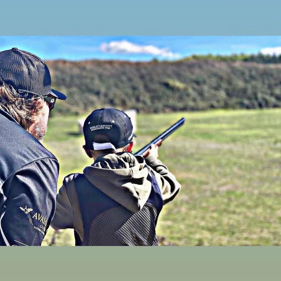 An instructor observing a young shooter aiming at a clay target.